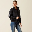 A woman in a black jacket with "ARIA" on the sleeve stands confidently. She wears blue jeans and has a relaxed expression, set against a plain background.
