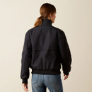A person with brown hair stands facing away, wearing a black jacket and blue jeans. The neutral background highlights a casual style.