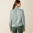A woman with a ponytail, dressed in a light blue long-sleeve top with "ARIAT" vertically on the back, and dark green leggings, stands facing away.