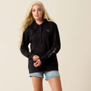 Young woman with long blonde hair in a black hoodie with logo, and "Ariat" on the sleeve, paired with frayed denim shorts, against a neutral background.