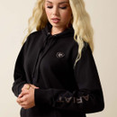 A person with long blonde hair wears a black hoodie with a small logo on the chest and "ARIAT" on the sleeve, standing against a plain background.