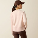 Woman wearing a pale pink sweatshirt with "Ariat International" logo, brown leggings, and a matching brown cap. She stands confidently, exuding a sporty, casual vibe.