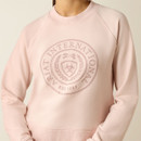 Woman wearing a light pink sweatshirt featuring an "ARIAT INTERNATIONAL" emblem with laurels and a shield. The mood is casual and stylish.
