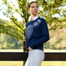 A woman in a blue sweatshirt with a crest logo stands outdoors by a wooden fence. She looks confidently to the side. Green trees and sunlight fill the background.