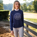 A person stands outdoors wearing a navy blue jumper with "Ariat International" text and emblem. They appear calm, with a vibrant green park backdrop.