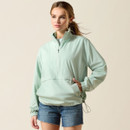 A woman in a light green windbreaker and denim shorts stands against a beige background. She has a relaxed pose and a confident expression.