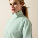 Young woman with a confident expression, wearing a light mint green high-collared jacket. Her hair is tied back, and she has small teal earrings.