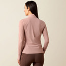 A woman with long dark hair, viewed from the back, wears a fitted, long-sleeve pink top and brown leggings. The calm, neutral background enhances the outfit's soft tones.