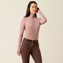 Woman wearing a light pink zip-up top and brown pants with a belt, standing with a neutral expression against a plain background.