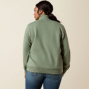 A woman with a ponytail wears a green sweatshirt and denim shorts, facing away. The neutral background emphasizes a casual, relaxed style.