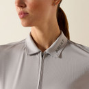 Close-up of a woman wearing a light gray zip-up jacket with "ARIAT" on the collar. Her hair is in a ponytail, and she projects a calm demeanour.