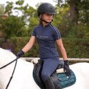 Young rider wearing a navy Aubrion Equestrian short-sleeve riding top and grey helmet, sitting on a white horse in an outdoor arena.