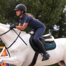 Action shot of rider jumping on a white horse, wearing a navy Aubrion Equestrian short-sleeve riding top, matching riding tights, gloves and black helmet.