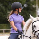 Side profile of a rider in a black riding helmet and lilac Aubrion Equestrian short-sleeve zip top on a white horse in an arena.