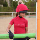 Young rider in a bright red Aubrion Equestrian short-sleeve riding top, wearing gloves and a red hat silk with pom pom, holding a green pole in an outdoor arena.