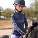 Young rider wearing a navy Aubrion Equestrian long-sleeve riding top and black helmet, seated on a black pony in an outdoor arena.