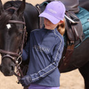 Child wearing a navy Aubrion Equestrian riding top and lilac Aubrion cap, standing beside a black horse with tack in an arena.