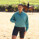 Woman modelling a teal hooded half-zip sweatshirt with kangaroo pocket, wearing black cycling shorts in an outdoor riding arena.