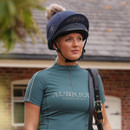 Rider wearing a navy Aubrion equestrian hat cover with faux fur pom pom over a riding helmet, outdoor lifestyle image.