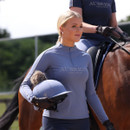 Blonde rider in a slate blue Aubrion Equestrian long-sleeve quarter-zip top holding a riding hat, with a horse and rider in the background.