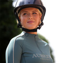 Close-up of a rider in a teal Aubrion Equestrian long sleeve quarter-zip top, wearing a glossy riding helmet outdoors.