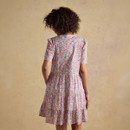 Back view of a woman in a pink floral tiered mini dress with short sleeves, relaxed fit summer dress, lightweight fabric, neutral beige backdrop.