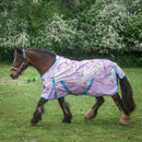 Side view of a bay cob walking on grass wearing a lilac unicorn-print turnout rug with turquoise straps, with flowering hedgerow in the background.
