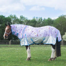 A horse stands side-on in a grassy paddock wearing a lilac unicorn-print turnout rug with a silver lower panel, turquoise binding and straps, and a neck cover.