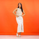 Full-length studio shot of a model wearing beige lace-up shoes with a cream sleeveless knit top and a white midi skirt against an orange backdrop.