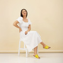 Lifestyle image of model seated on a white stool wearing a white dress and bright yellow crossover strap mule sandals against a beige backdrop.