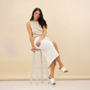 Lifestyle image of a model seated on a stool wearing a beige sleeveless knit top, white midi skirt and white crossover strap mule sandals against a beige backdrop.
