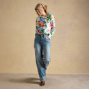 Full-length studio shot of a model wearing a white long-sleeve floral top with blue jeans and a headscarf, posed against a beige backdrop.
