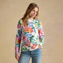 Model wearing a white long-sleeve top with a colourful painted floral print, styled with blue jeans against a beige studio backdrop.