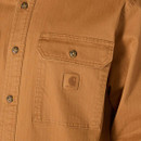 Close-up of the left chest pocket on the tan Carhartt shirt jacket, showing the flap button and Carhartt logo patch.
