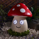 Smiling mushroom-shaped garden light with a red and white spotted cap and illuminated LED eyes, glowing at night among decorative stones.
