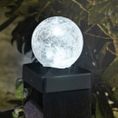 Close-up of a crackle-glass ball garden light glowing white, mounted on a dark fence post surrounded by foliage at night.