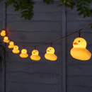 String of glowing yellow rubber duck fairy lights hanging outdoors against a garden fence at night.