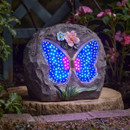 Stone-look garden rock with a glowing LED butterfly design in blue and pink, decorated with small flowers, sitting on a patio in a garden.