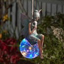 Hanging fairy garden ornament showing a seated fairy with wings perched on a glowing multicolour crackle-glass ball, suspended among plants at night.