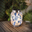 Solar glass lantern with a metal handle and stainless-steel top, glowing warm white through blue-and-white speckled mosaic glass, sitting on a wooden outdoor table with greenery in the background.