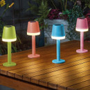Set of four small solar table lamps with built-in top panels and glowing white bands, in green, coral, blue, and pink, arranged on a wooden outdoor table with plants in the background.
