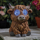 Highland cow garden ornament with shaggy brown fur and horns, sitting on a stone slab with bright blue LED “goggle” eyes glowing against leafy plants.
