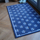 Blue doormat with white floral patterns and a thin border, placed on a wooden floor near a window. Light blue slippers rest at the corner, conveying a cosy feel.