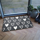 A grey doormat with white heart patterns sits on a wooden floor beside a glass door. Nearby are potted plants and black boots, creating a cosy, inviting atmosphere.
