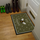 Doormat with "Bee Happy" text, featuring a bee design on a green polka dot background. Nearby are vibrant green and gray boots on wooden floor.