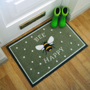 Doormat with a bee graphic and the phrase "Bee Happy" on a green background, placed near a white door. Bright green boots add a cheerful touch.