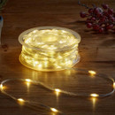 Coiled reel of clear string lights on a wooden table, glowing warm white, with a short section unspooled in front and decorative red berries in the top-right background.