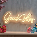 Warm white neon “Good Vibes” sign in cursive script mounted on a textured wall above a shelf with flowers and small paper umbrellas.