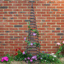 Tall brown spiral garden obelisk trellis standing in a flower bed against a red brick wall, with purple and pink flowers growing around it.