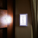 Wall-mounted white LED night light with an on/off switch glowing softly beside a wooden bed frame and curtain.
